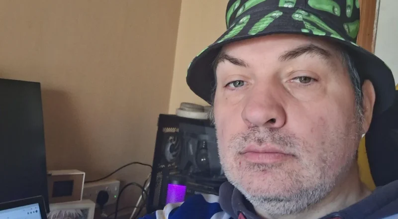 St Richard's Hospice Man with a short grey beard takes a selfie wearing a black bucket hat with cartoon pickles on it. He is sat in an office environment with beige/yellow walls and a computer in front of him.