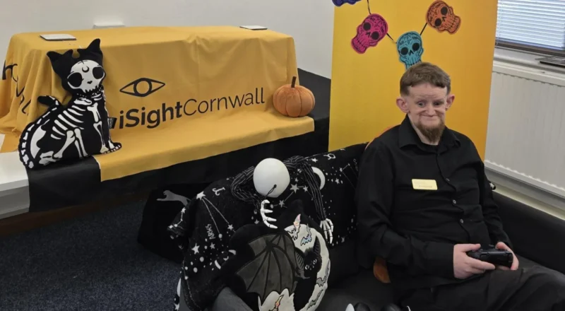 iSightCornwall Dominic Hall sitting on a chair. He is wearing a black shirt and black trousers. He is holding a black gaming console. The chair he is sat on has a black blanket with a bat and moon on it. Behind him is a pillow of a cat with a skeleton. There is an iSightCornwall logo behind him and a pumpkin pillow. The walls around him are white and the floor has grey carpet.