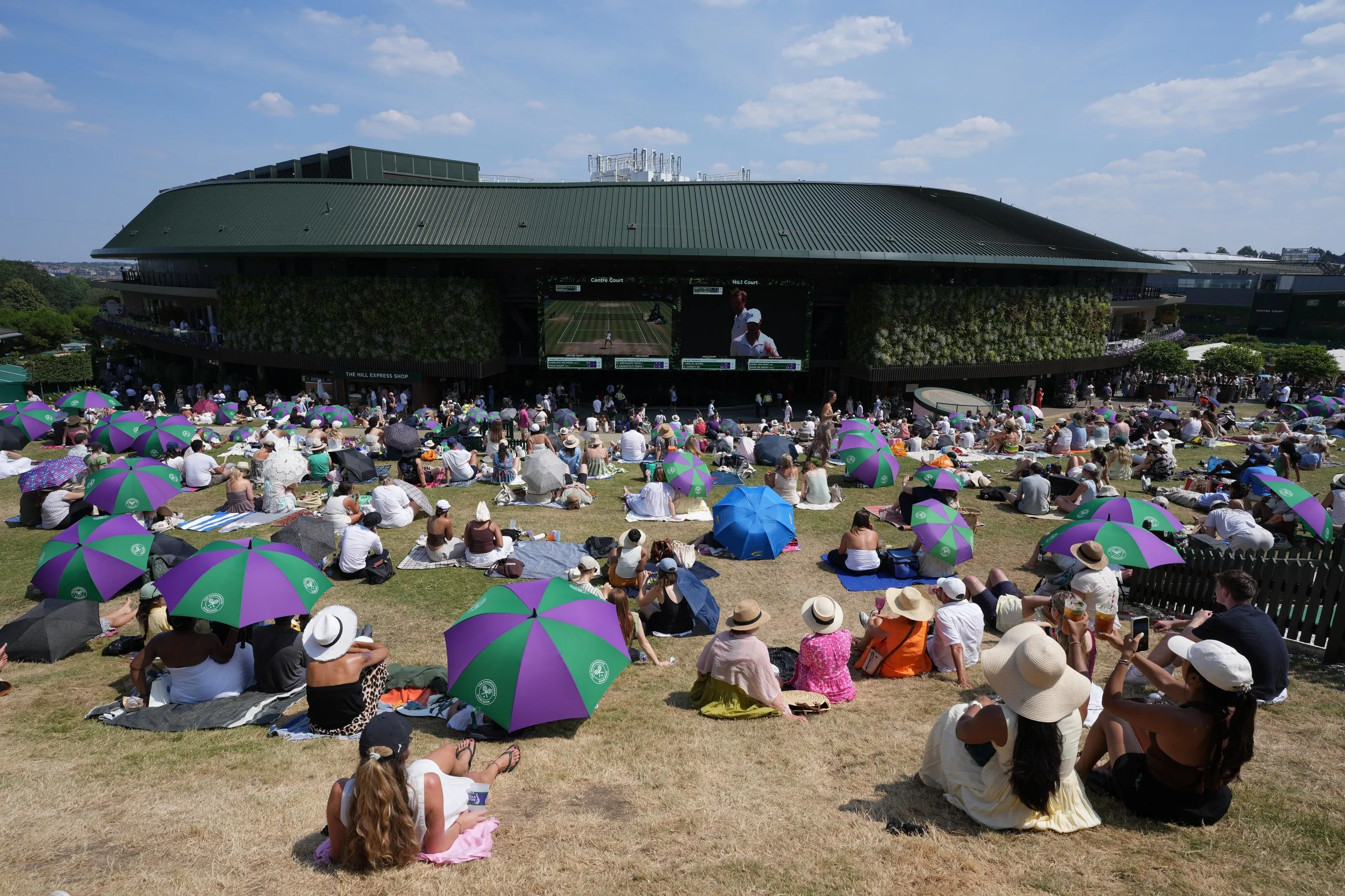 Fans sit on the weathered grass of Henman Hill at Wimbledon, some sheltering from the sunshine under umbrellas, watching tennis on large video screens